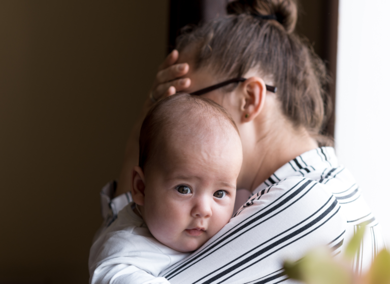 Image of a mother holding her baby while holding her head. Representing what postpartum depression symptoms feel like. There is hope though with postpartum and perinatal therapy in Edmonton and Sherwood Park, AB.