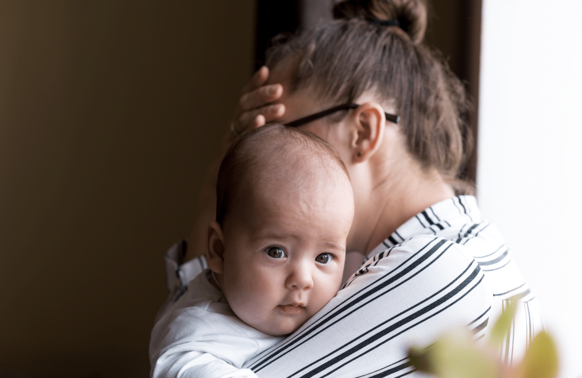 Image of a mother holding her baby while holding her head. Representing what postpartum depression symptoms feel like. There is hope though with postpartum and perinatal therapy in Edmonton and Sherwood Park, AB.